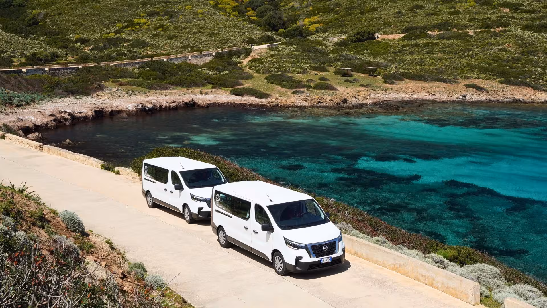 Two white minivans parked by the turquoise waters of Asinara island, highlighting the Stintino tour route.