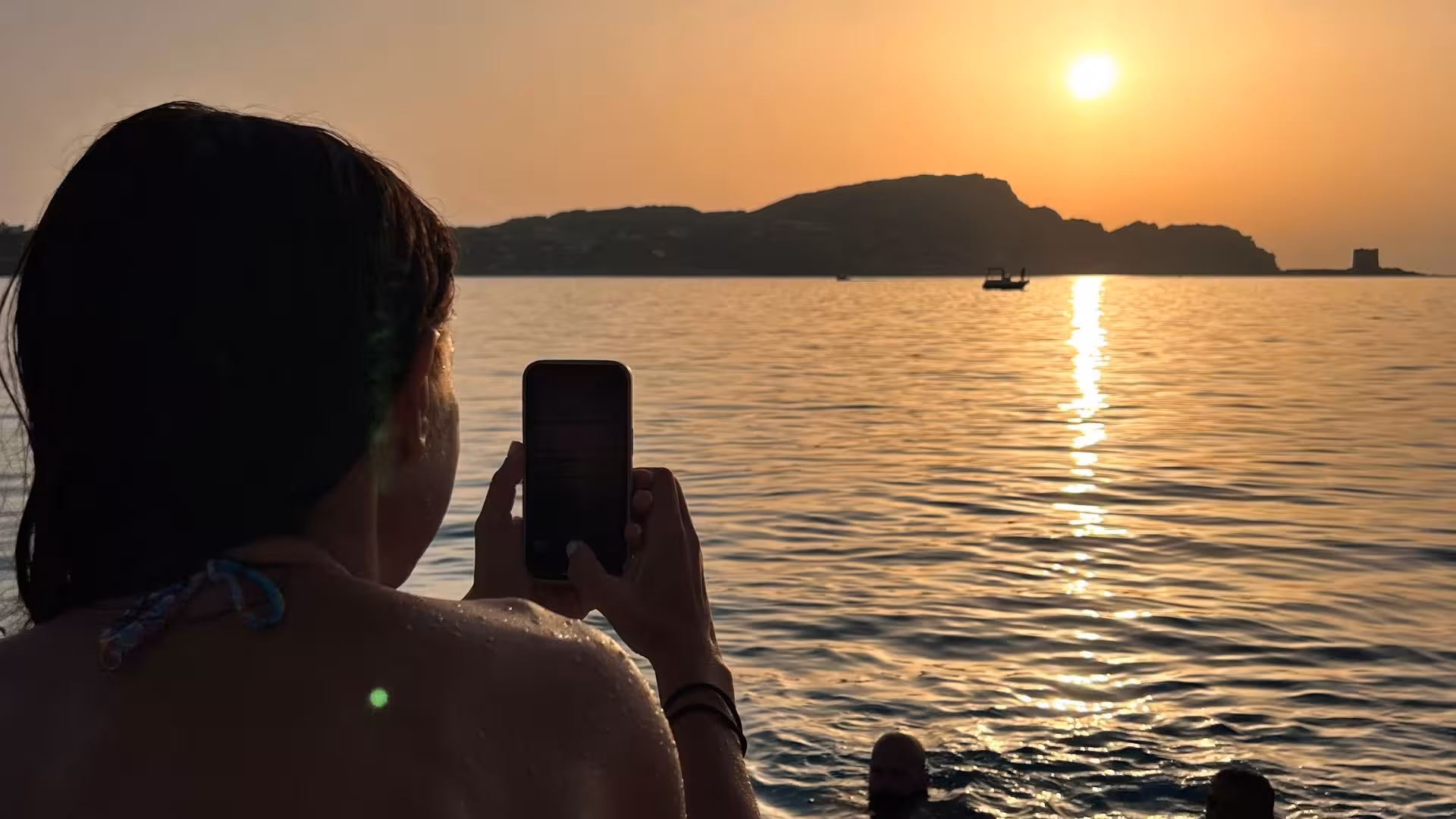 Person photographing a stunning sunset over the Gulf of Asinara from a catamaran with distant boats.