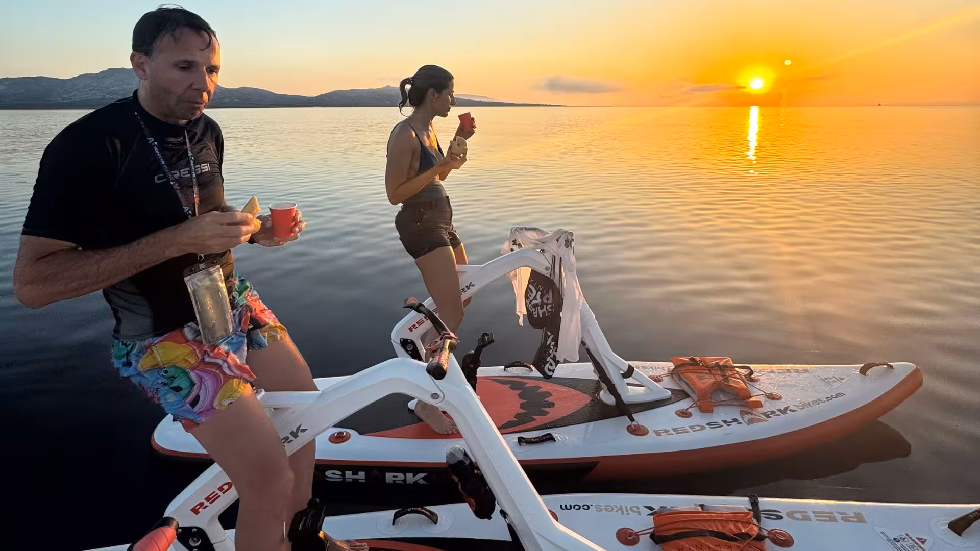 Two people relaxing on water bikes with snacks while watching the sunset at La Pelosa, Stintino.