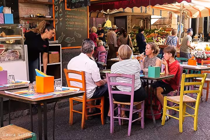 People enjoy dining at a lively outdoor street market in Catania, surrounded by colorful chairs and bustling stalls.