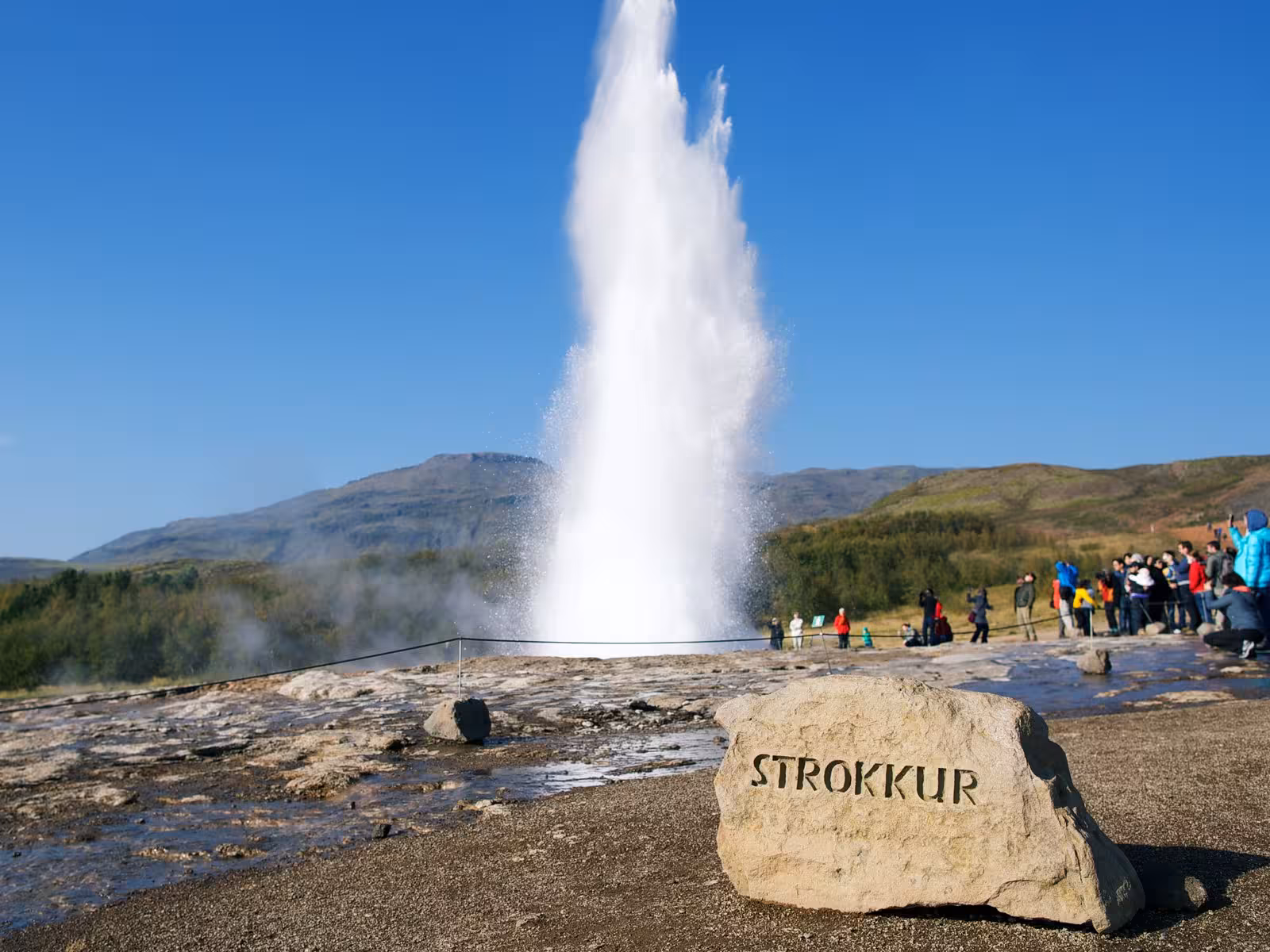 Tourists watch the Strokkur geyser erupt against a clear blue sky in South Iceland's Golden Circle.