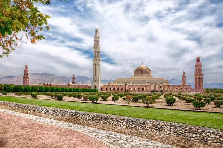 Panoramic view of the Sultan Qaboos Grand Mosque in Muscat, featuring its majestic minarets and lush gardens under a cloudy sky.