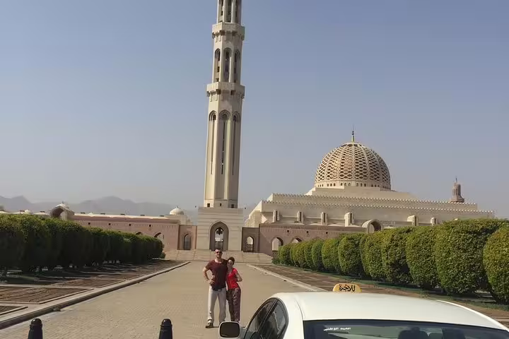 Visitors admire the architectural beauty of Sultan Qaboos Grand Mosque in Muscat under a clear blue sky.
