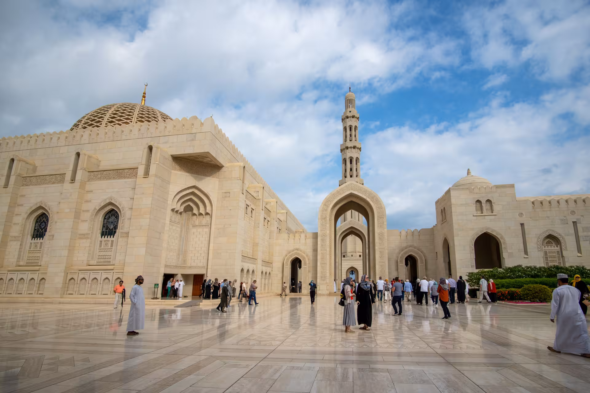 Visitors exploring the marble courtyard and grand minaret of Sultan Qaboos Grand Mosque during a private Muscat city tour