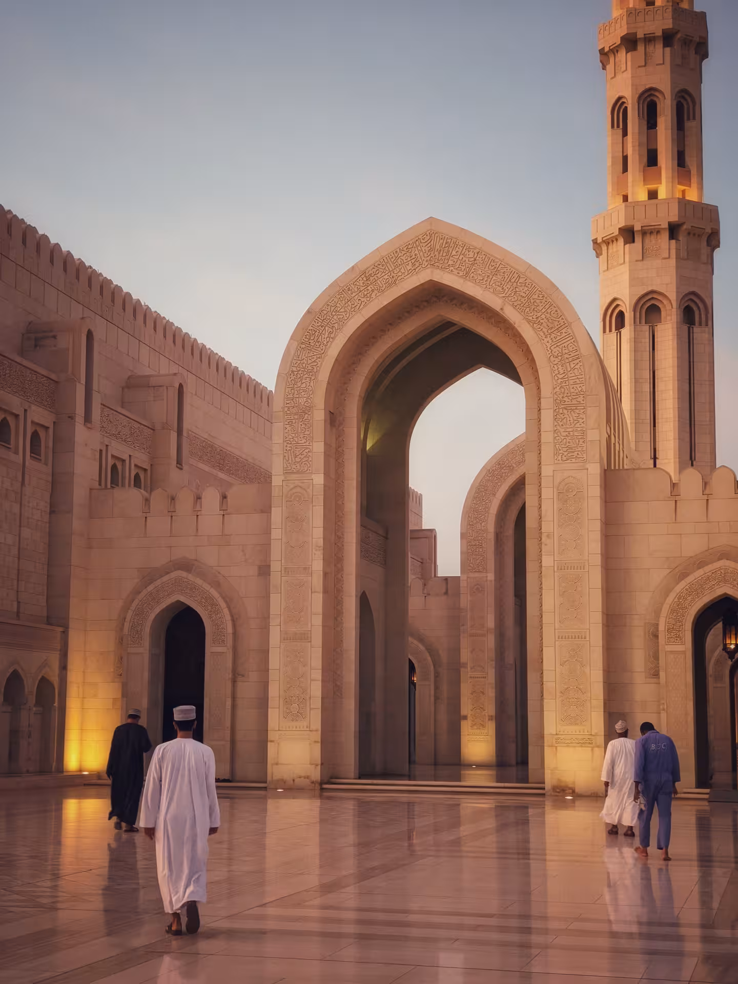 Visitors walk through the illuminated marble arches of Sultan Qaboos Grand Mosque at dusk on a private Muscat city tour