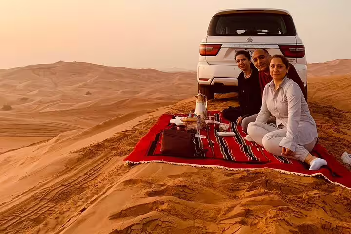 Tourists enjoy a sunrise picnic on a red blanket in the Dubai desert during a private safari with a parked SUV.