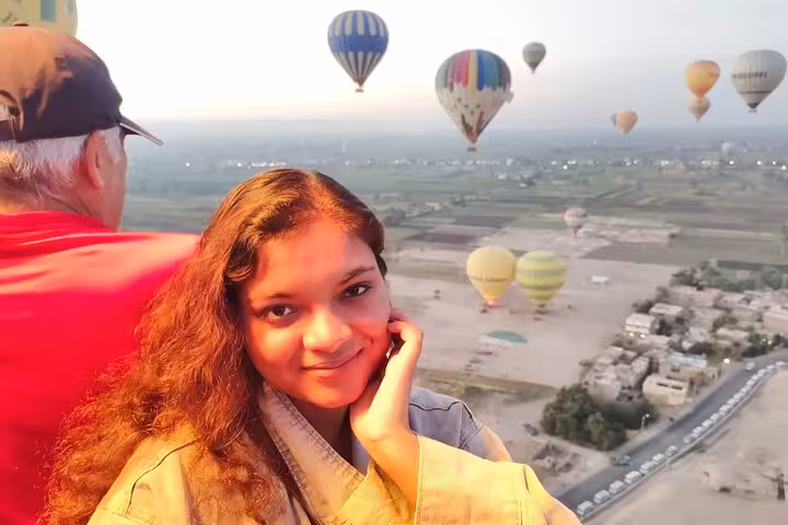 Smiling traveler enjoys a sunrise hot air balloon ride over Luxor West Bank, with scenic views and colorful balloons in the background.