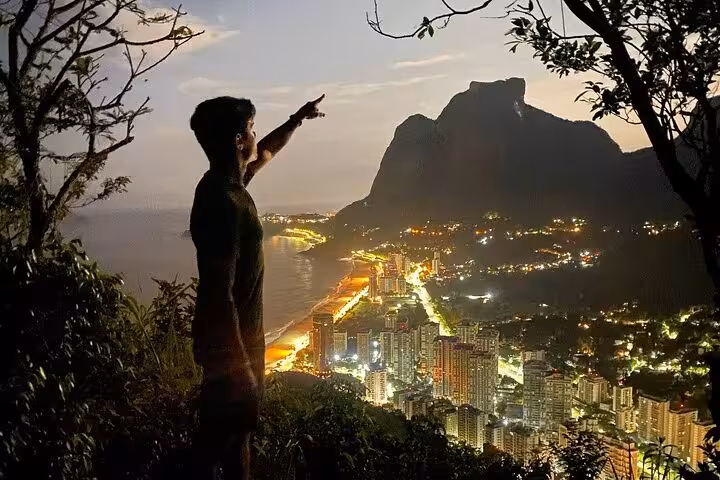 Person pointing at sunrise view over Two Brothers Mountain and illuminated cityscape in Rio de Janeiro.