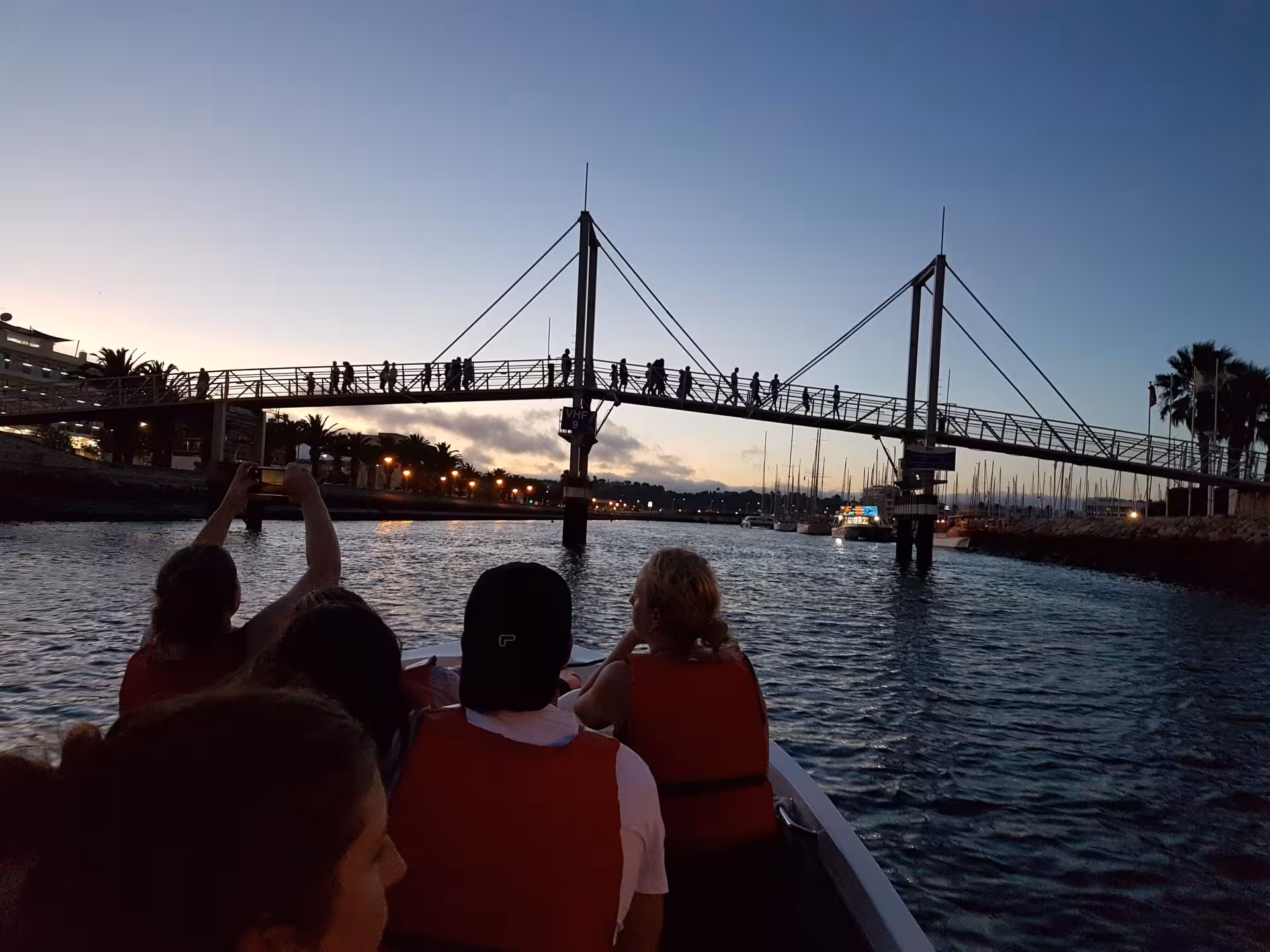 Travellers in life jackets enjoy a Lagos sunset boat trip under the marina footbridge as lights glow along Ponta da Piedade