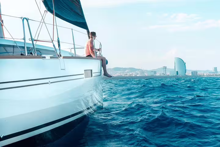 Person relaxing on a sailboat during a sunset cruise near Barcelona, with city skyline and blue sea in the background.