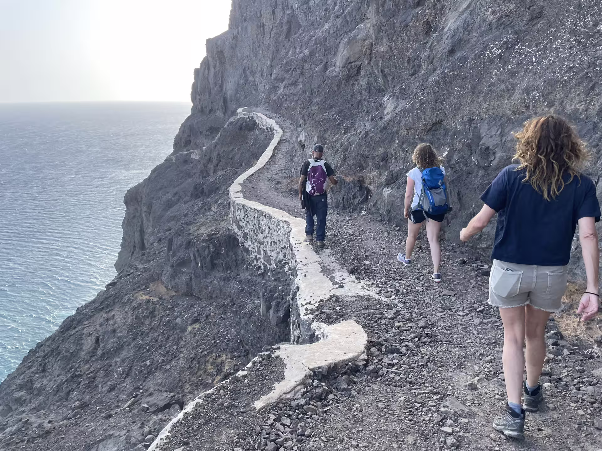 Hikers trek along a rugged coastal path towards Lighthouse Dona Amélia during a sunset hike, with ocean views and rocky cliffs.