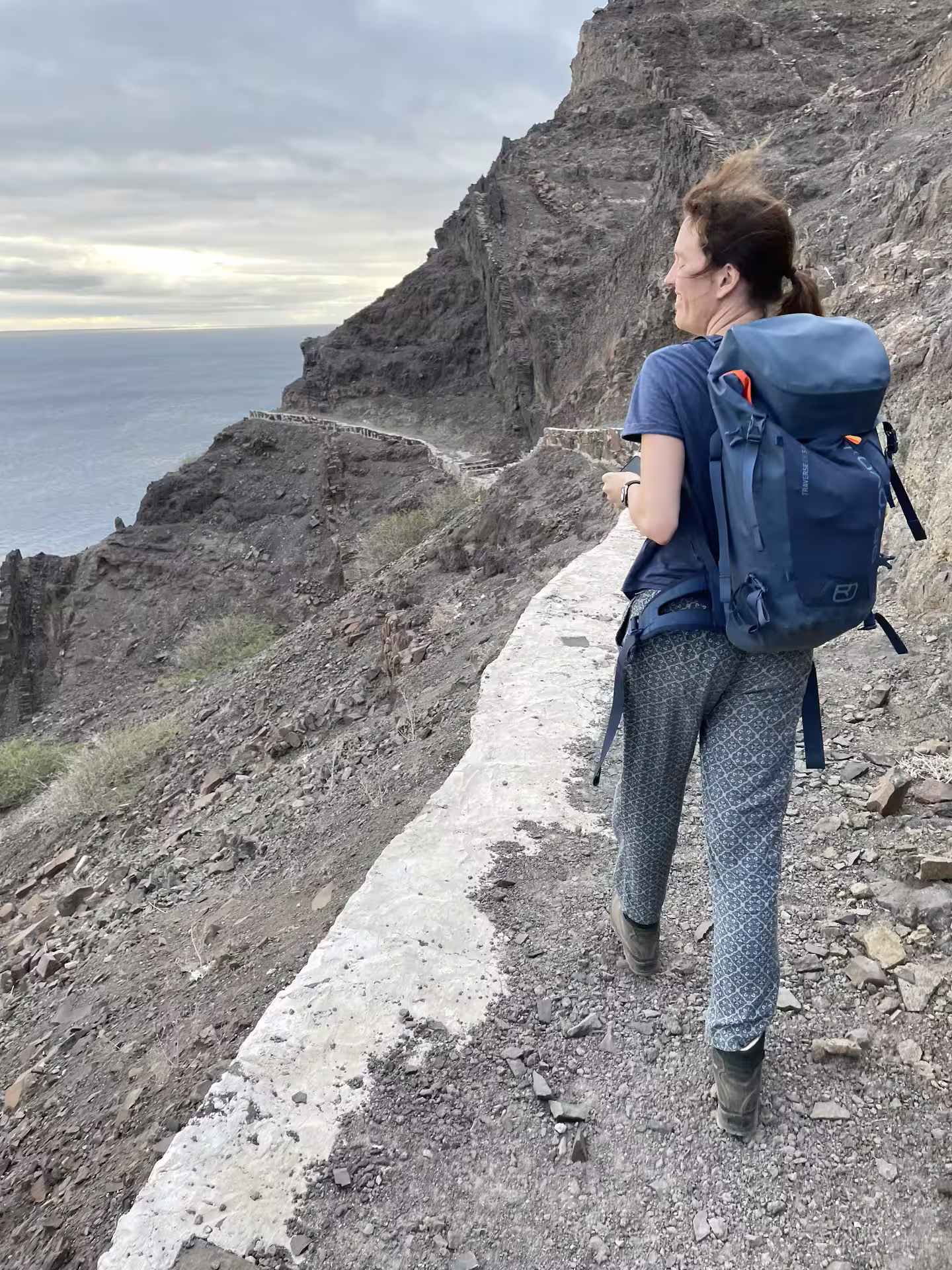 Hiker with a backpack enjoying a scenic sunset view on a rocky trail to Lighthouse Dona Amélia, overlooking the ocean.