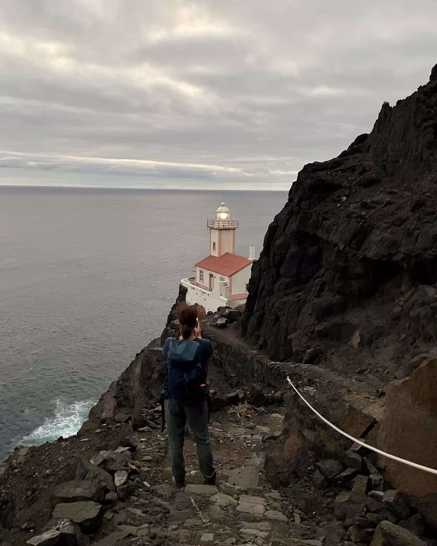 Hiker photographs the iconic Lighthouse Dona Amélia at sunset, perched on a rugged cliffside overlooking the ocean.