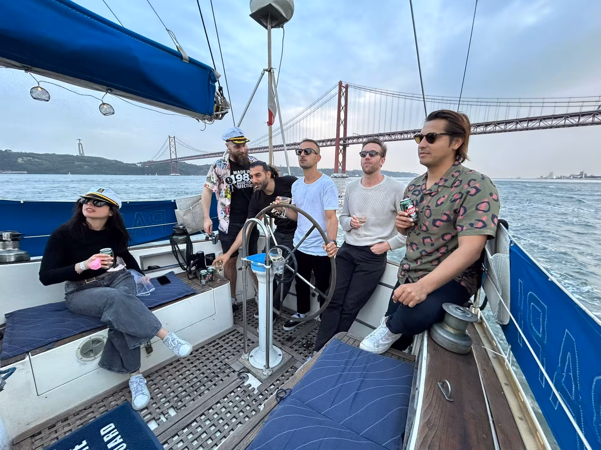 Friends relaxing with drinks on a shared sunset sailboat tour beneath Lisbon’s 25 de Abril Bridge