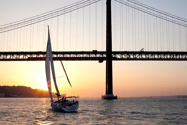 Sailboat gliding under the iconic bridge on the Tagus River at sunset, offering picturesque views of Lisbon.