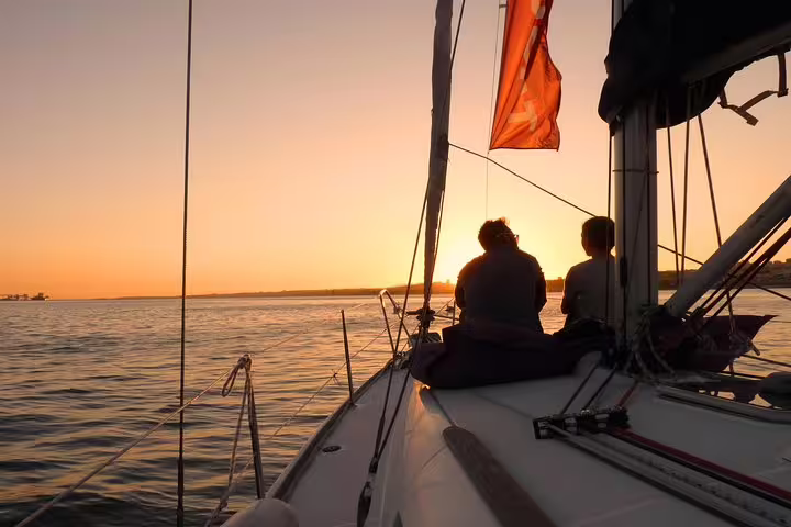 Two people enjoying a serene sunset on a sailboat cruising the Tagus River, with a warm orange glow in the sky.