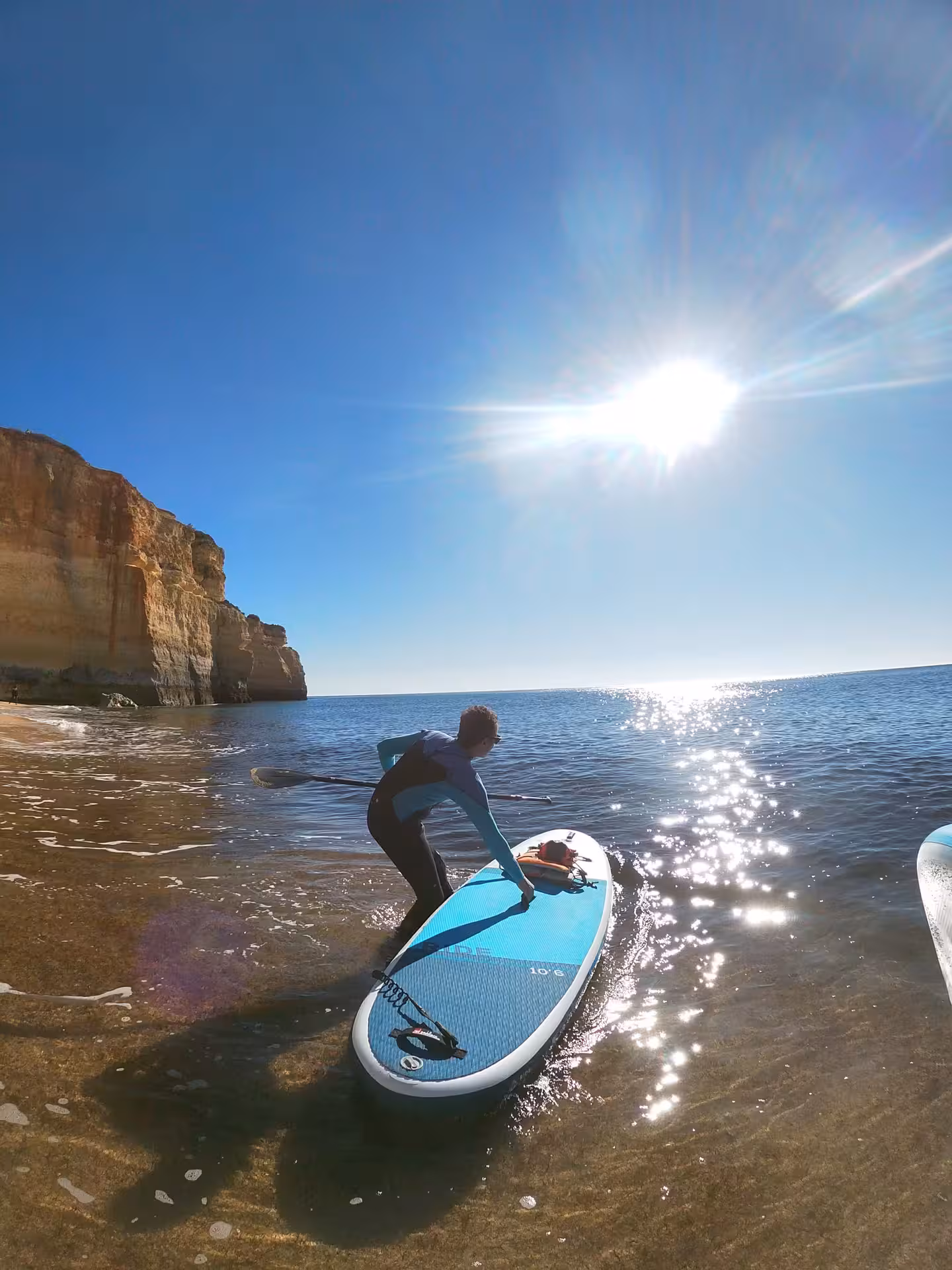 Paddleboarder launching SUP into clear Atlantic waters below golden Algarve cliffs on a sunny Portugal beach day