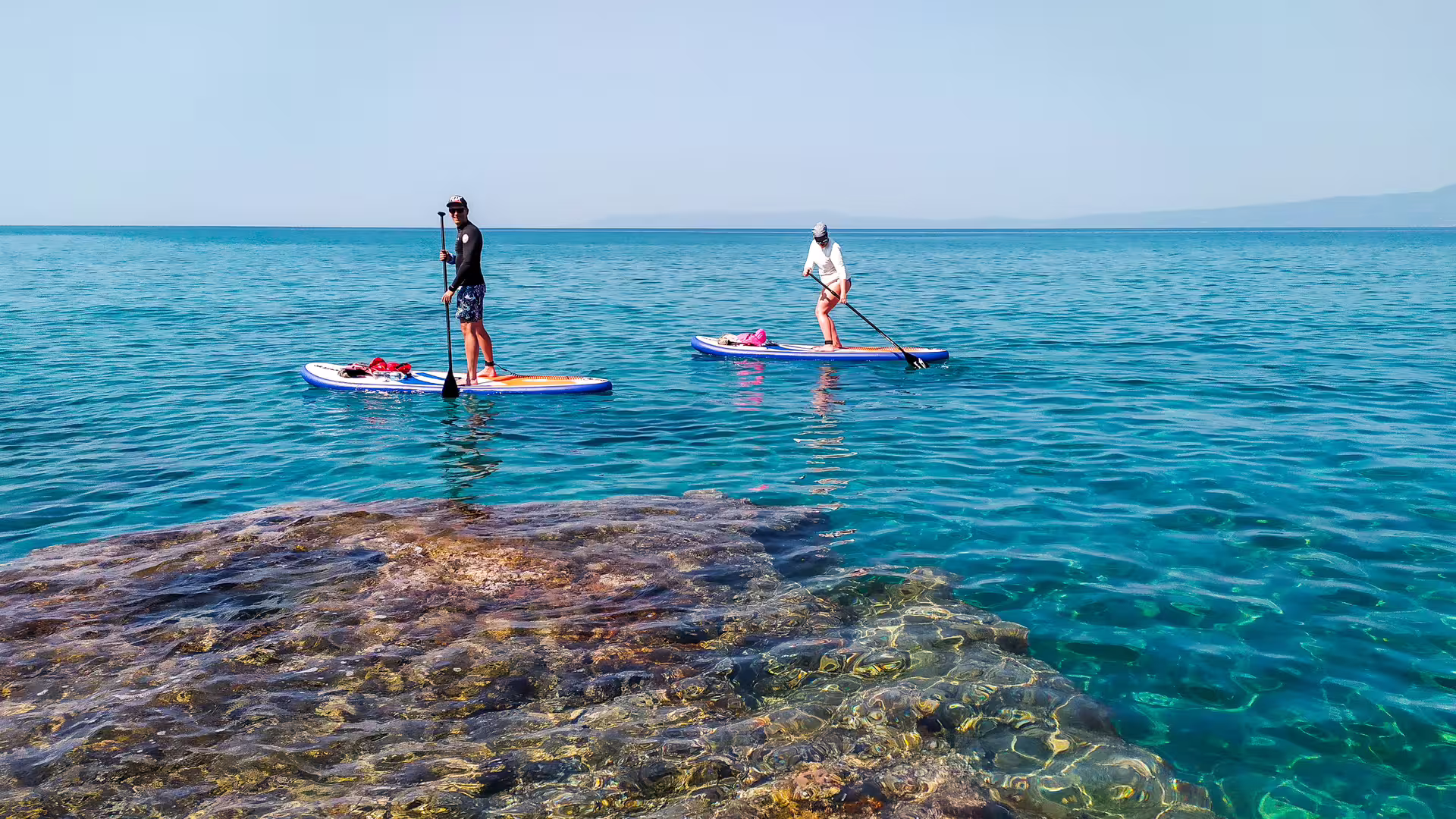 Two paddleboarders on calm turquoise sea during SUP tour in Kalamata, Messinia, Greece near rocky shore
