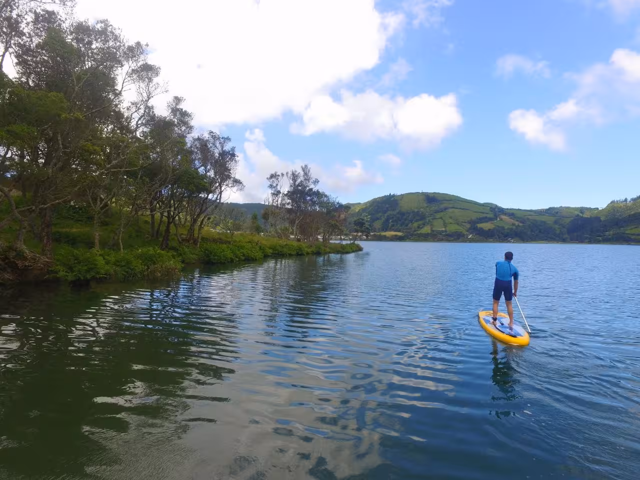 Man on stand up paddle board exploring tranquil Sete Cidades lake shoreline with green hills and blue sky in the Azores
