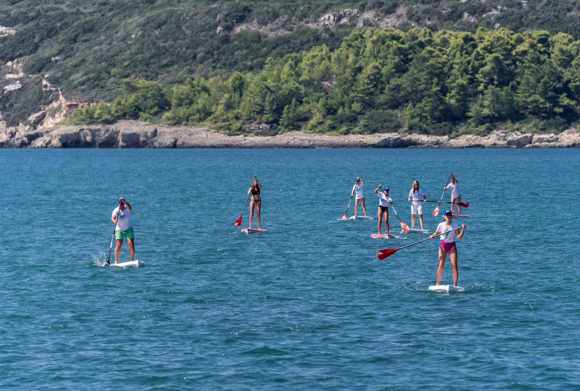 Group stand-up paddleboarding on turquoise Voidokilia Bay, Messinia, with pine-covered cliffs in the background