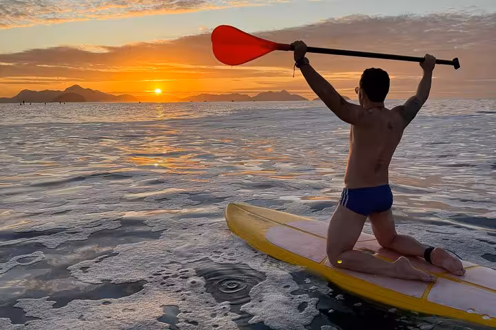 Experience sunrise paddleboarding at Copacabana with a vibrant orange sky and a paddler raising a paddle triumphantly.