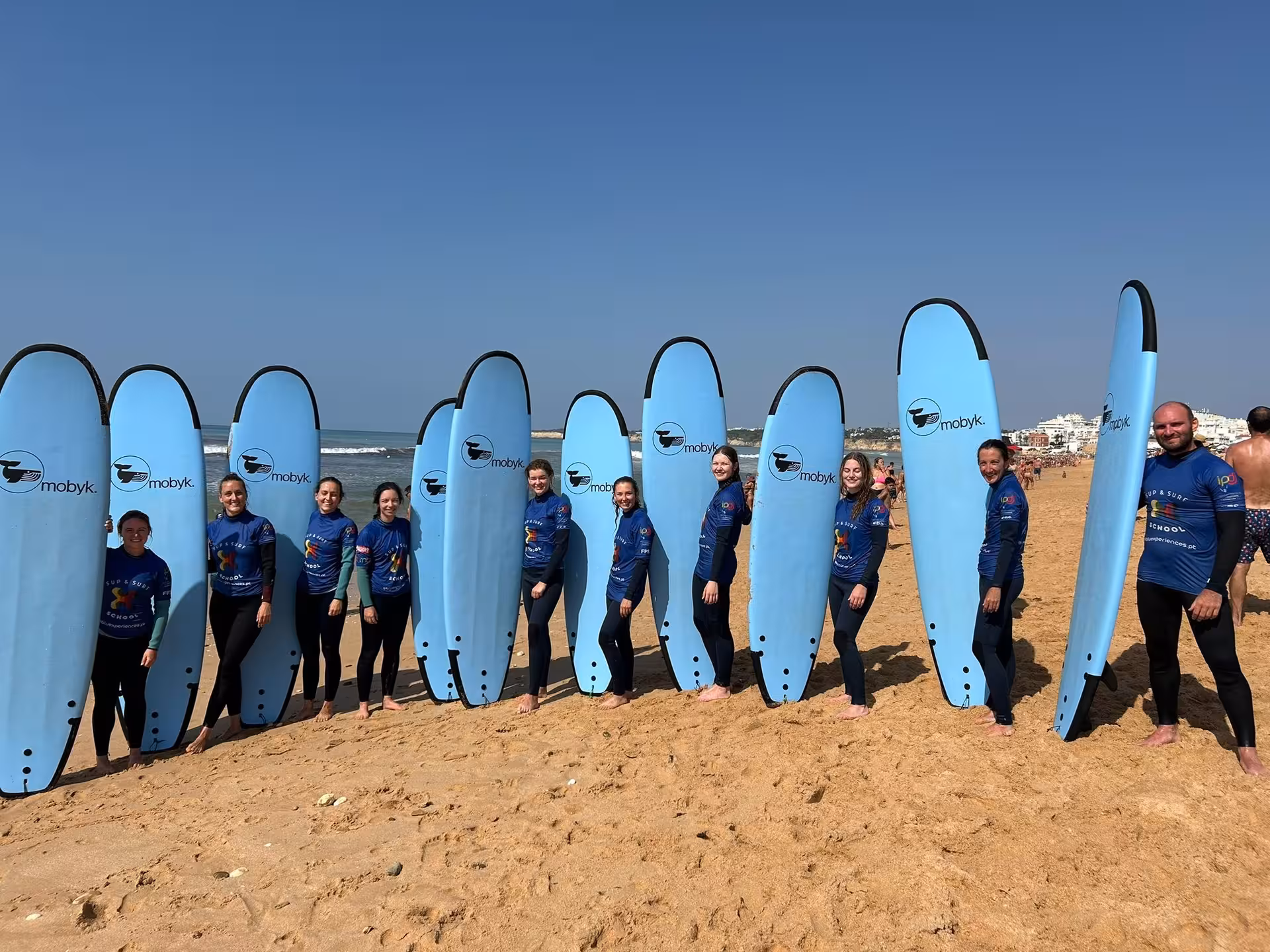 Happy surf school group posing with blue beginner boards on golden sand after a fun surf clinic session