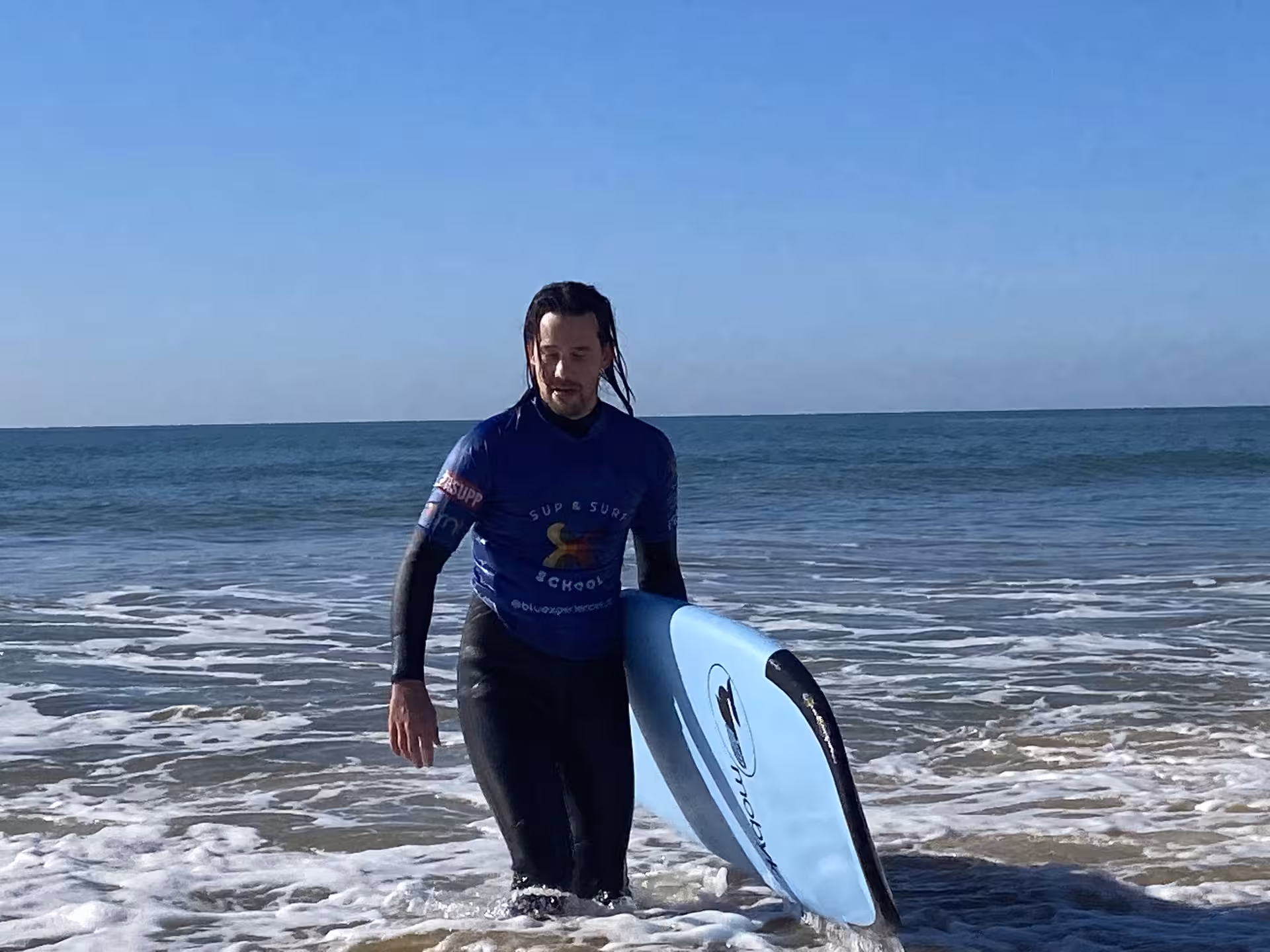 Surf student in wetsuit walking out of the sea with a blue foam board after a surf experience class
