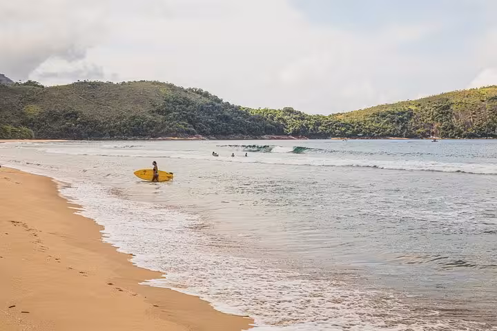 Surfer heading into the gentle waves of Sono Beach, surrounded by lush green hills, perfect for water sports enthusiasts.