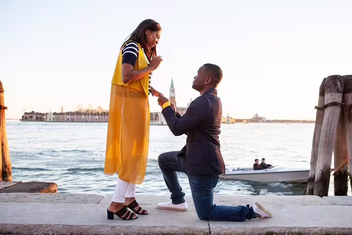 Surprise proposal on Venetian lagoon at golden hour, groom on one knee as photographer captures engagement moment