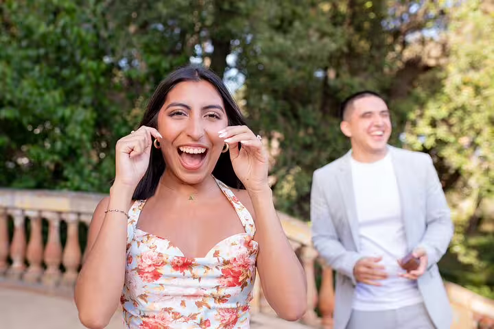 Surprised bride-to-be reacts during Barcelona proposal photo shoot in a sunny park with partner behind