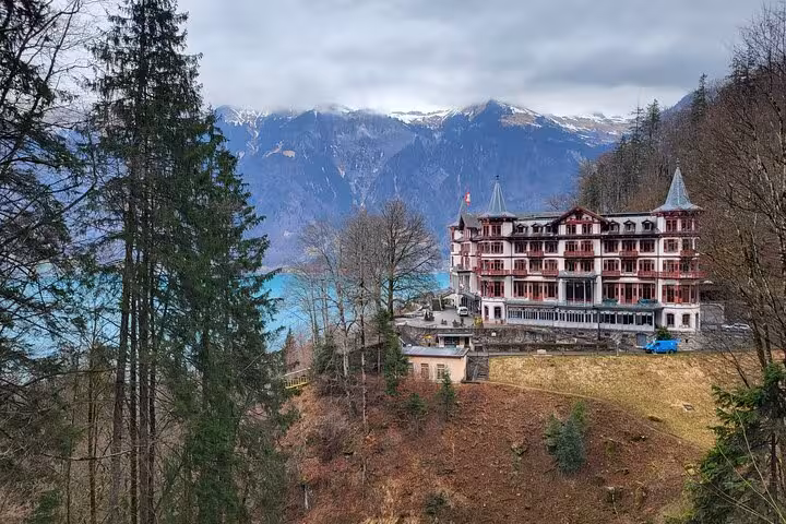 Historic Swiss hotel nestled among trees with majestic alpine lake and mountain backdrop, ideal for a serene getaway.