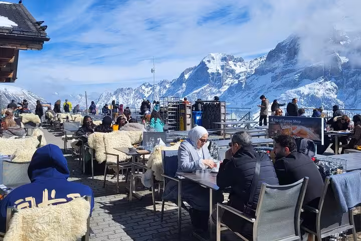 Tourists enjoying a scenic outdoor café with breathtaking views of snow-covered Swiss Alps on a sunny day.