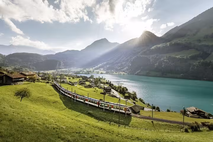 Scenic view of a Swiss train traveling along a turquoise lake surrounded by majestic mountains and green fields.