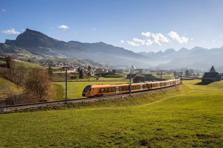 Orange Swiss train gliding through lush green valleys with mountain backdrop on scenic express route.