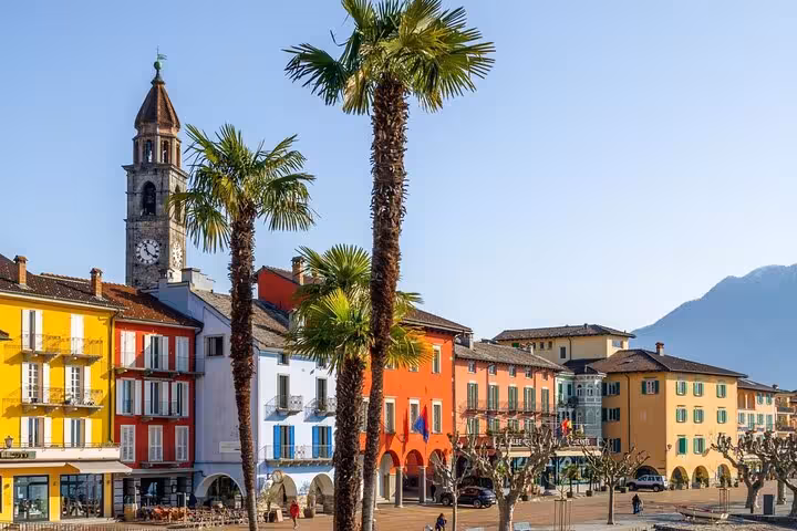 Scenic Swiss lakeside town with colorful buildings, palm trees, and a historic clock tower.