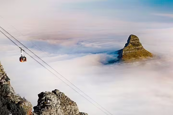 Aerial view of a cable car ascending Table Mountain through clouds, with Lion's Head peak in the background.