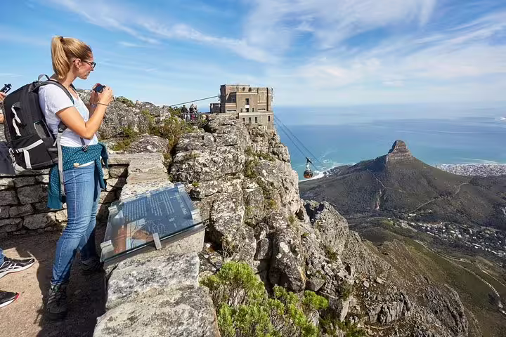 Tourist photographing panoramic views from Table Mountain's summit, featuring cable car and iconic Cape Town landscapes.