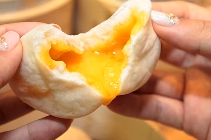 Hands holding a freshly steamed bun with a rich, golden custard filling oozing out at Taipei Hungzhou Xiaolongbao.
