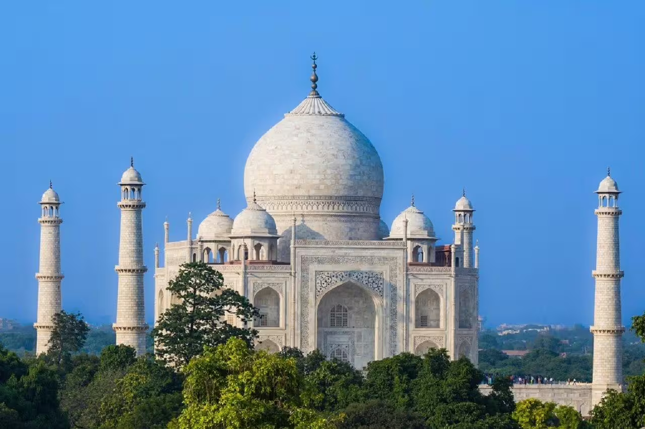 Iconic Taj Mahal framed by lush greenery, showcasing its stunning marble architecture against a clear blue sky.