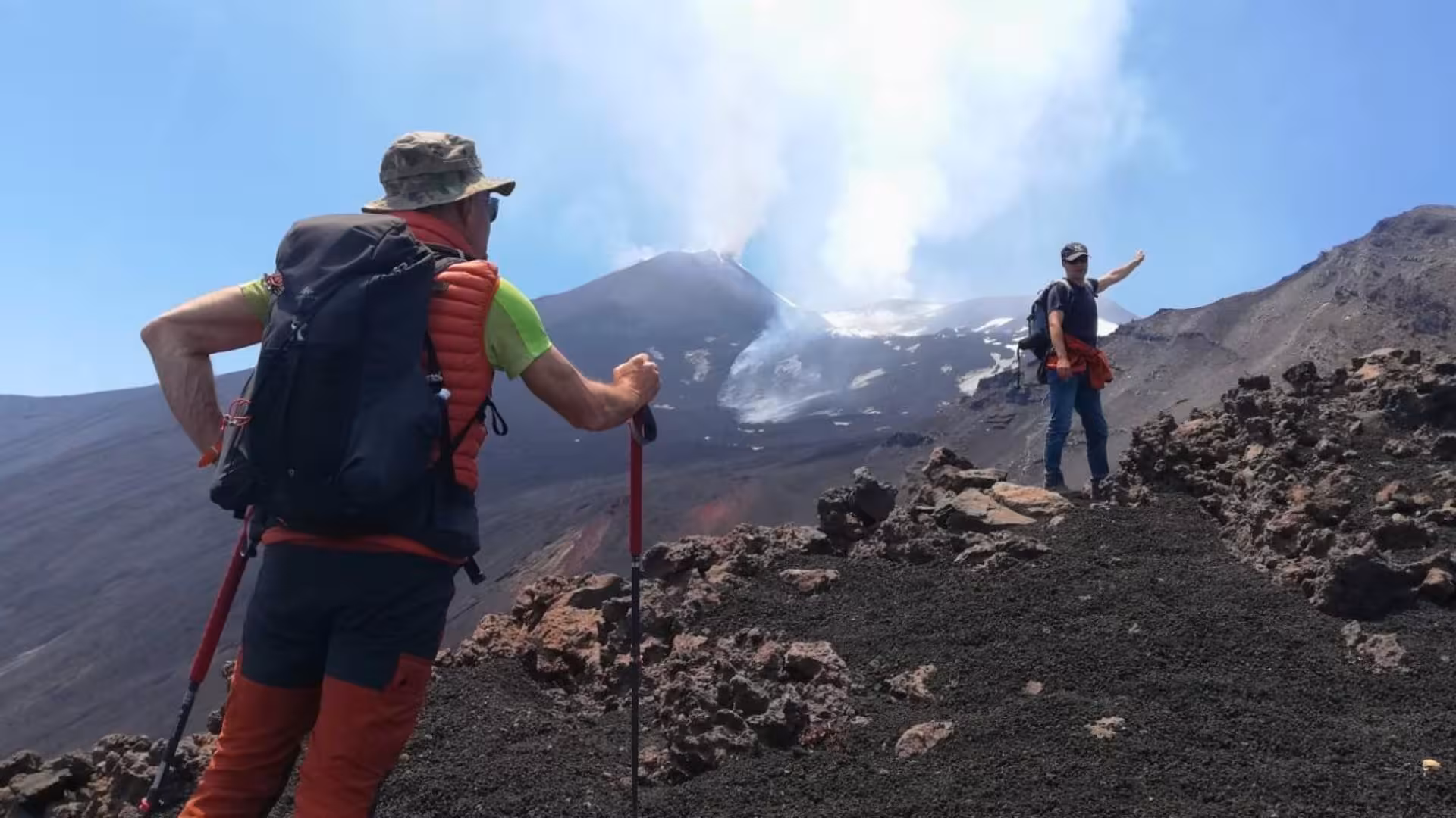 Two trekkers on rocky terrain observing smoke rising from Mount Etna's crater, highlighting adventure and nature.