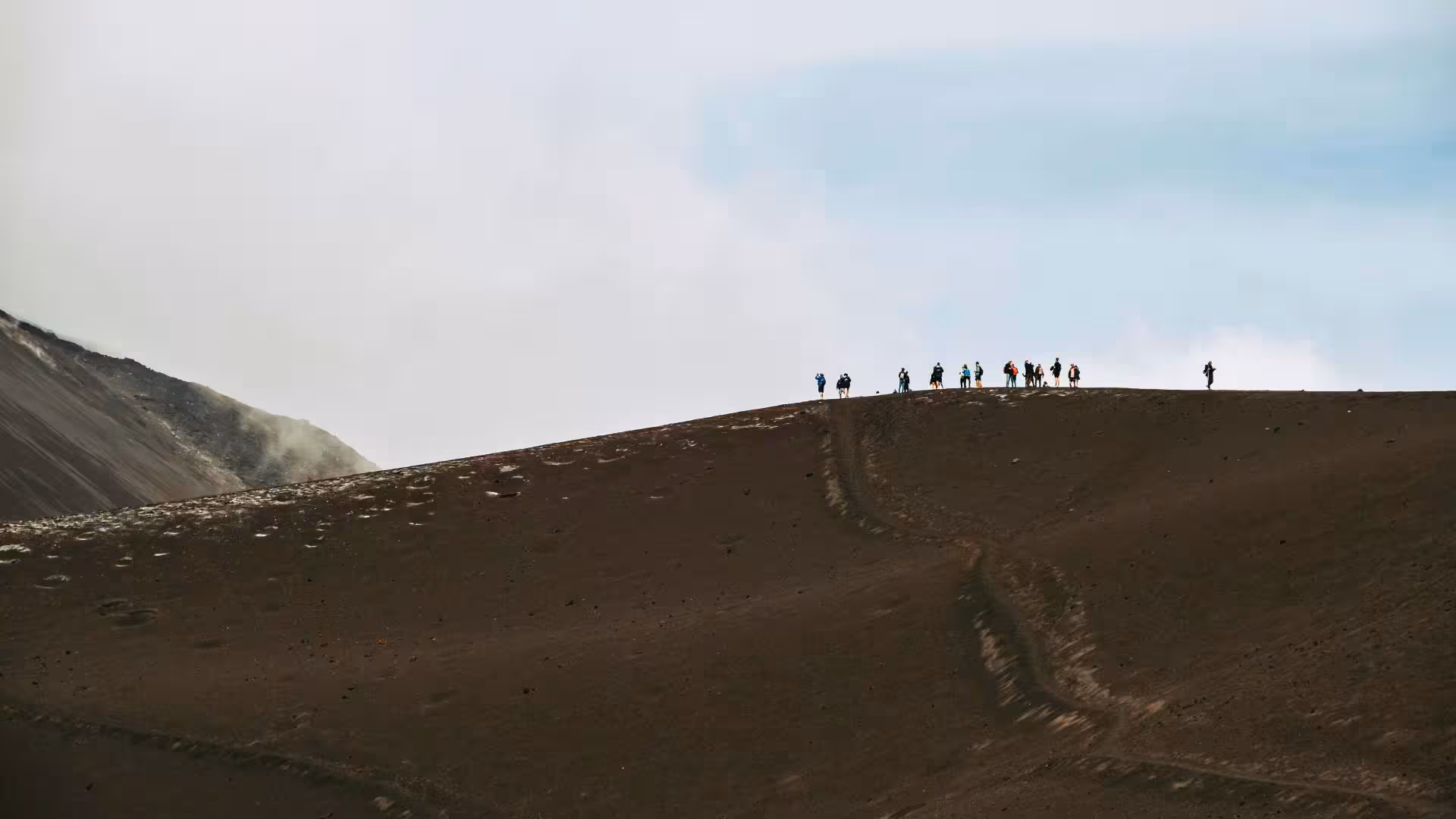 Silhouetted hikers walking along the ridge of Mount Etna under a vast sky, showcasing the dramatic volcanic landscape.