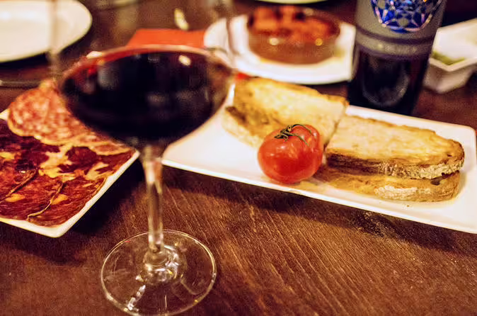 Close-up of a tapas spread with cured meats, toasted bread, tomato, and a glass of red wine on a wooden table.