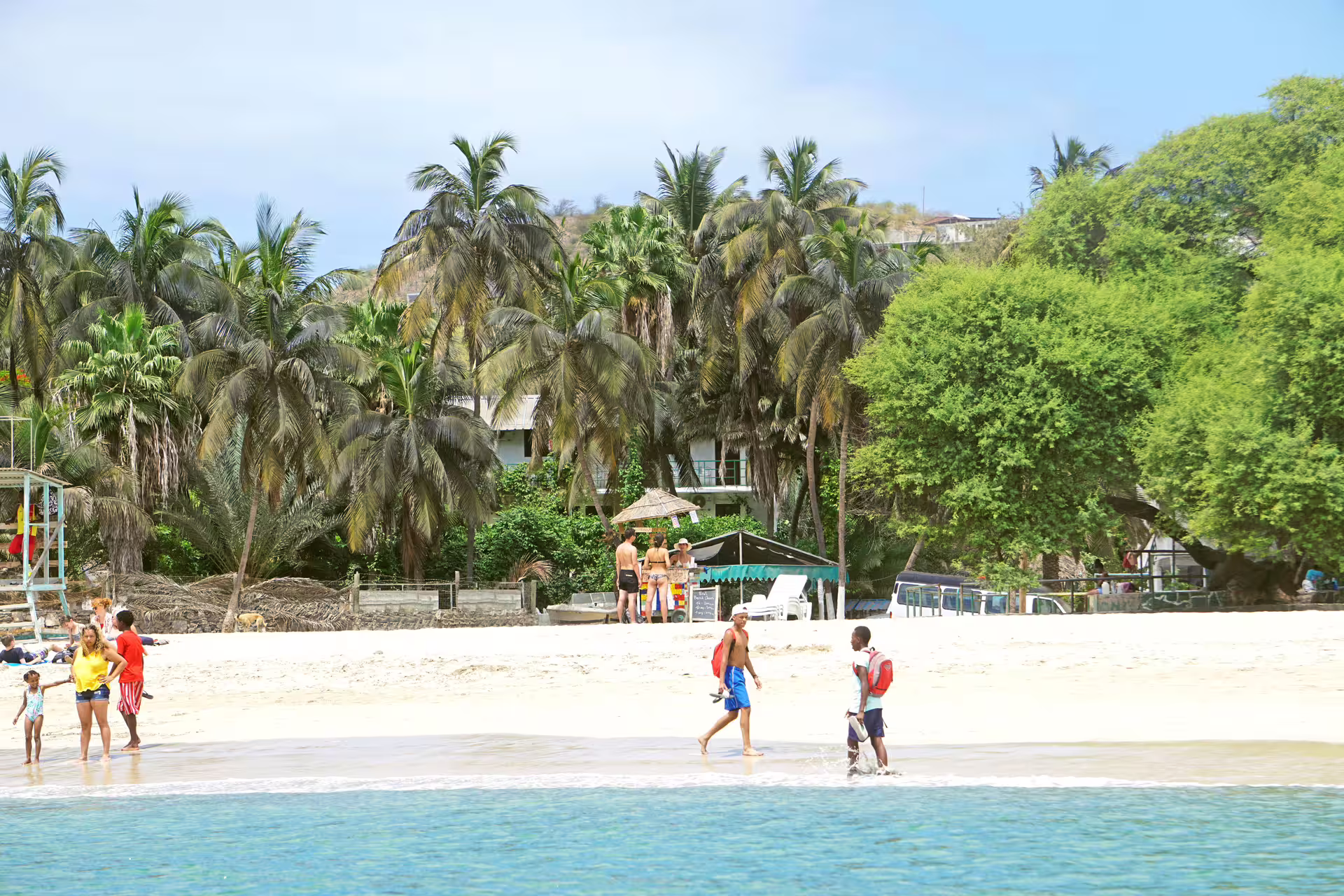 Families and tourists stroll along the sandy shores of Tarrafal Beach, surrounded by vibrant palm trees.