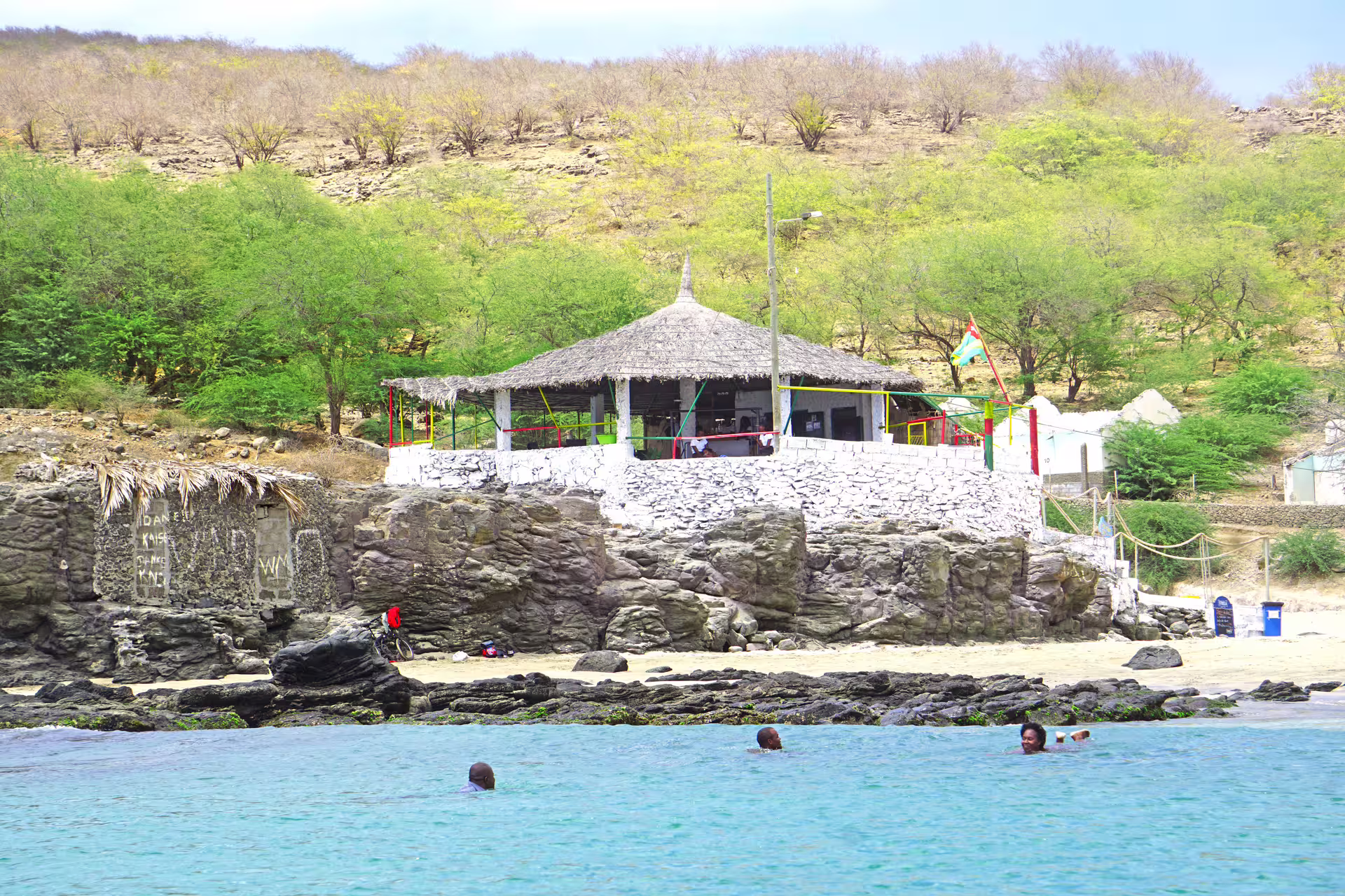 Scenic view of Tarrafal Beach with a rustic hut, lush greenery, and swimmers enjoying the turquoise waters.