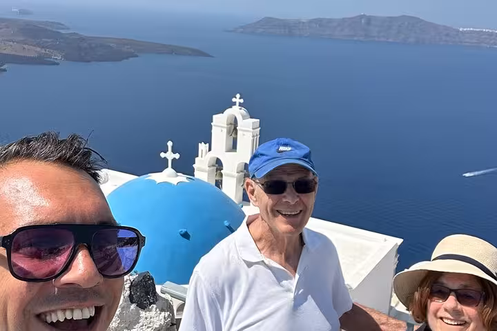 Couple and guide selfie by Oia blue dome overlooking Santorini caldera on Thera day tour with lunch and wine tasting
