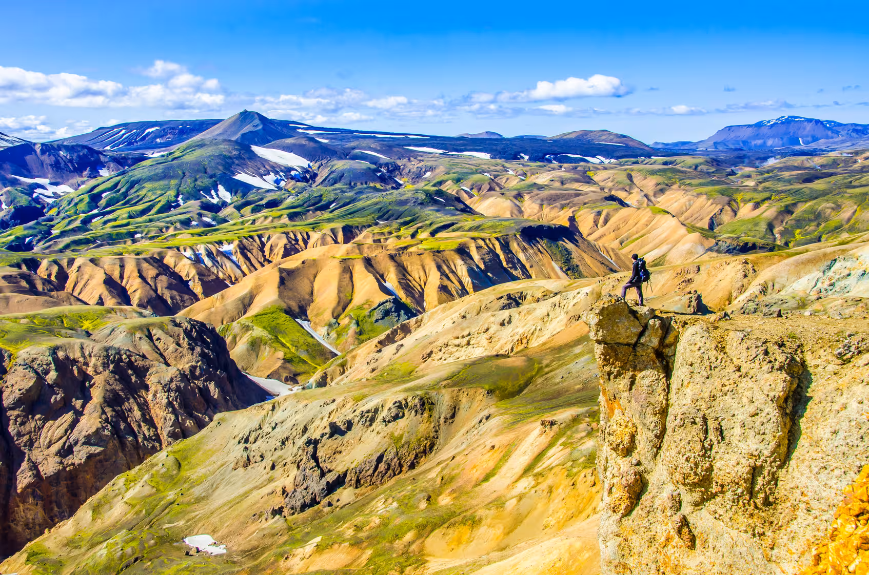 Adventurer stands atop a rocky cliff overlooking the colorful, expansive Thorsmörk Glacier Valley under a clear blue sky.