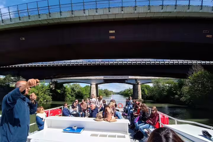 Tour group relaxing on open-deck boat cruising the Tiber River from Rome to Ostia Antica under modern bridges and green banks