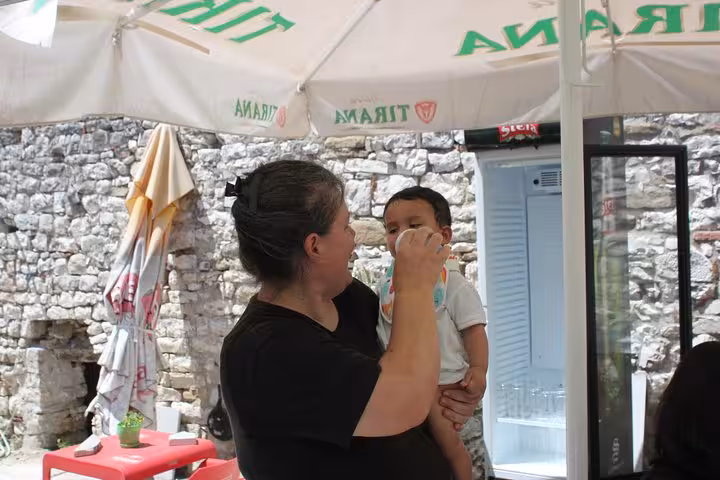 A woman holding a baby under a Tirana umbrella in a rustic courtyard setting in Berat during the city tour.