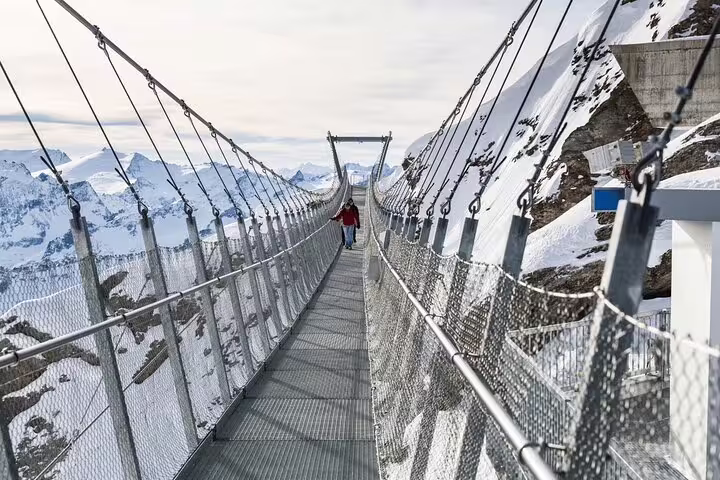 A person walks across the dramatic Titlis suspension bridge, surrounded by breathtaking snowy mountain vistas.