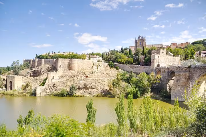 Scenic view of Toledo's historic walls and Alcantara Bridge along the Tagus River on a sunny day.